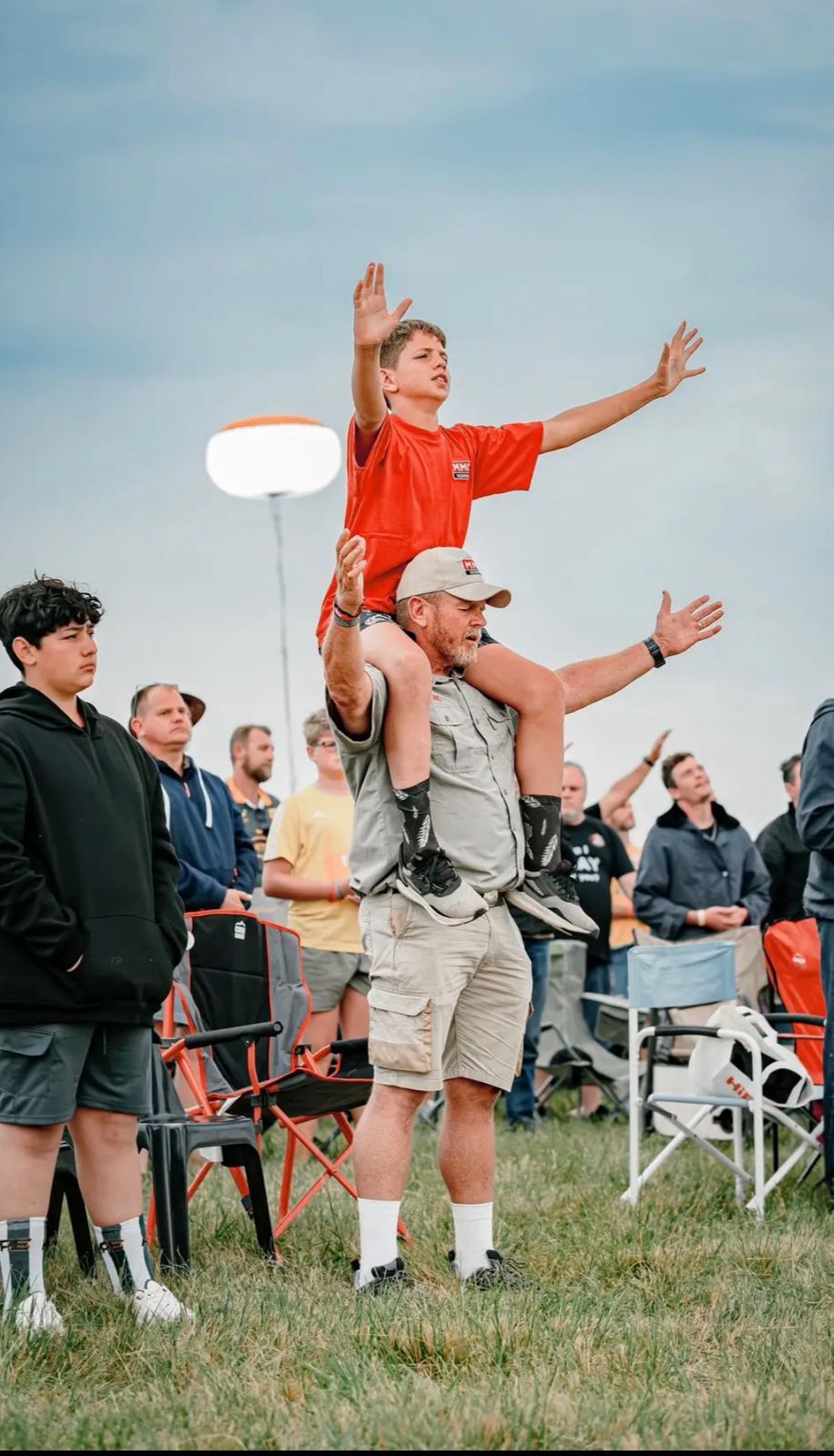 A boy on his father's shoulders with arms raised in worship